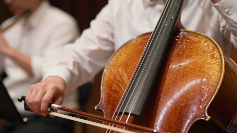 A close-up photograph of cello strings and a bow mid-motion. The focus is sharp on the resin dust and wood grain, with a background of other musicians in soft ice white shirts.