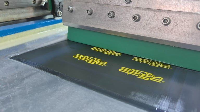 Automatic screen printing machine applying yellow ink onto a black textile panel with a green squeegee.