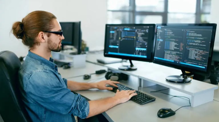 A young programmering is programming at a desk with 2 computer screens