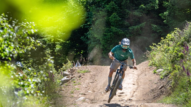 A mountain biker wearing a helmet and goggles jumps over a dirt trail in a green forest.
