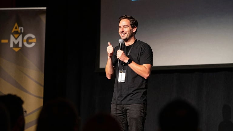 An MC on a stage at a North American / US tech conference. The lighting is professional with dark charcoal and muted gold stage accents. The MC is holding a microphone and smiling at the audience.