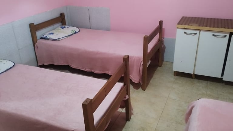 Dormitory room with wooden twin beds, pink bedding, and a small white cabinet.