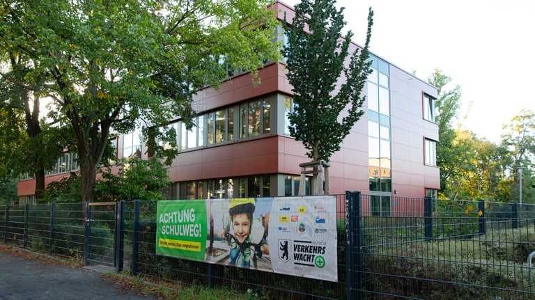 Foto von einerm roten Schulgebäude, an dem Zaun davor ist ein Plakat zur Schulwegsicherheit.