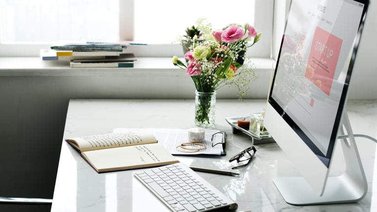 A bright workspace with keyboard, monitor, mouse, desk accessories, and flowers in a vase next to a window.