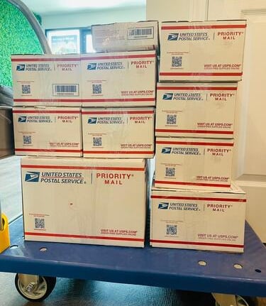 Stack of USPS Priority Mail boxes on a blue shipping cart ready for delivery or pickup.