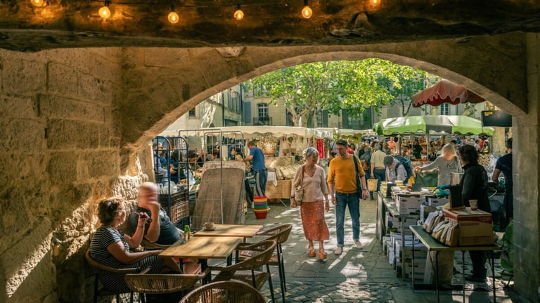 Crowded outdoor farmers market in a French village seen through a stone archway with café seating.