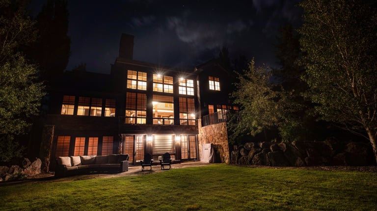 Luxury mountain home with glowing windows at night featuring a stone patio and grassy lawn.