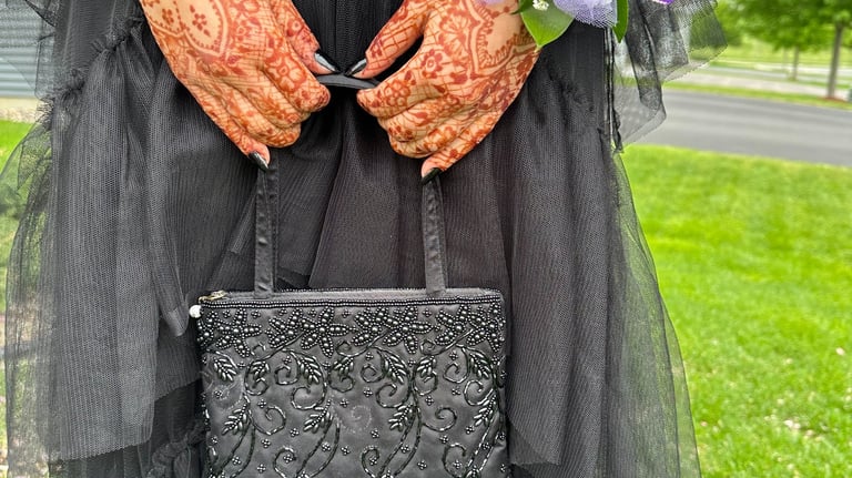 Close up of two hands with henna on them, holding a purse strap, wearing a black dress & corsage