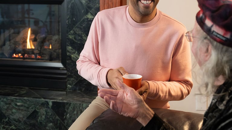 Smiling young man talking to a senior friend by a cozy fireplace while holding a coffee mug.