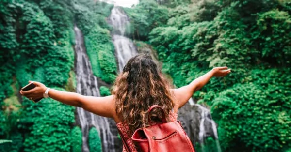 A female traveler with a red backpack stands with arms wide open before a lush tropical waterfall.