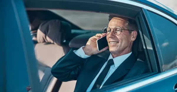 Smiling businessman in a suit talking on a smartphone while riding in the back of a luxury car.