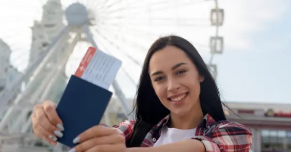 A smiling young woman holding a passport and flight boarding pass in front of a white ferris wheel.