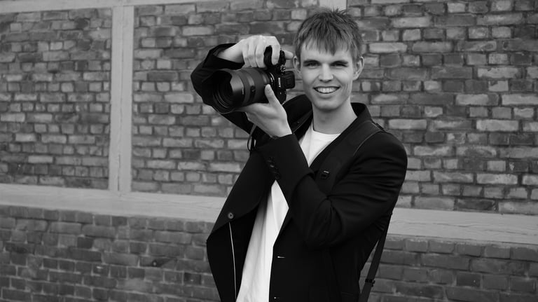 Schwarz-Weiß-Aufnahme von Joel Pingel mit Kamera vor einer Ziegelwand. Eventfotografie: Just Perspectives.