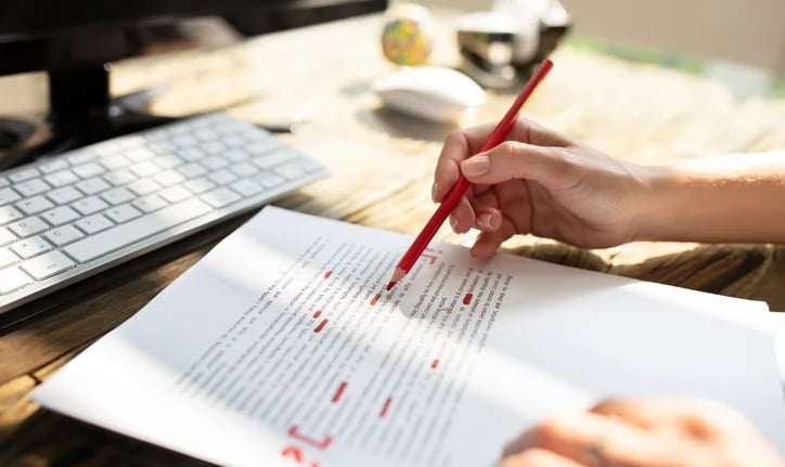 a person proofreading and correcting a document with a red pencil, computer in the background
