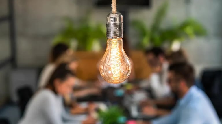 A focused lit and hanging lightbulb with individuals sitting at a table blurred in the background.
