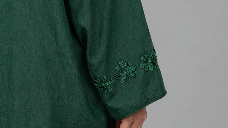 A close-up photography of a forest green linen abaya sleeve featuring delicate hand-stitched floral embroidery. The fabric has a natural, premium texture. Soft-focus background with English color name warm grey. Middle Eastern / Gulf style.