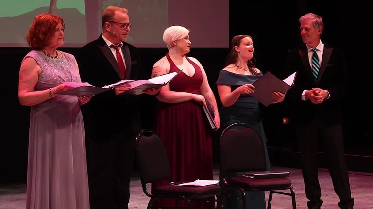 Five singers in formal attire performing on stage; four watch the soloist as she performs.