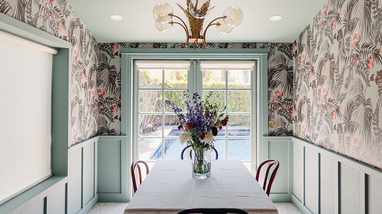 Modern dining room featuring tropical floral wallpaper, sage green wainscoting, and a view of a backyard pool.