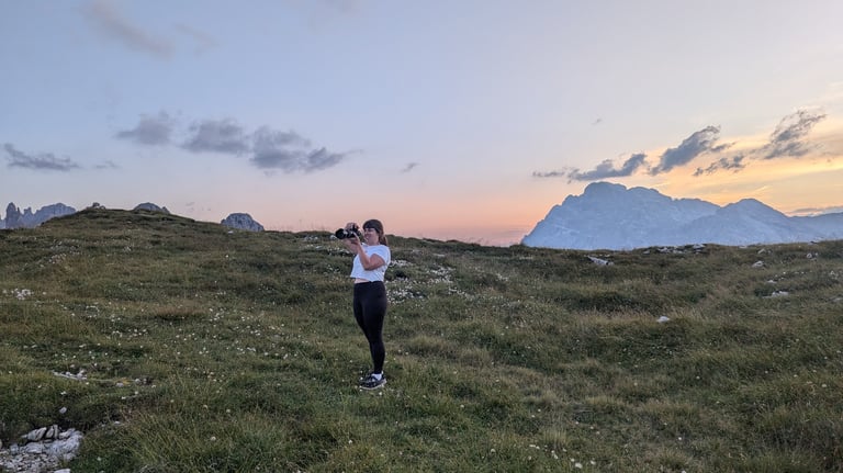 Laura stands on a green field at sunset taking a photo. The Italian Dolomites are in the background