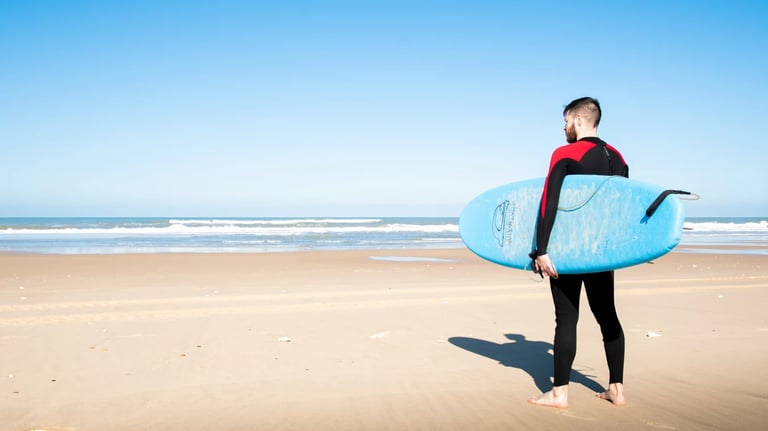 En homme en combinaison de surf, avec une planche bleu sur une plage