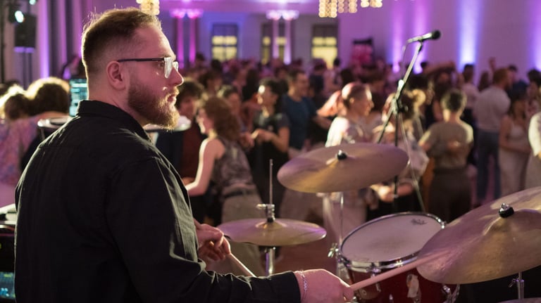 A bearded male drummer plays a live drum set at a crowded indoor party with purple lighting.