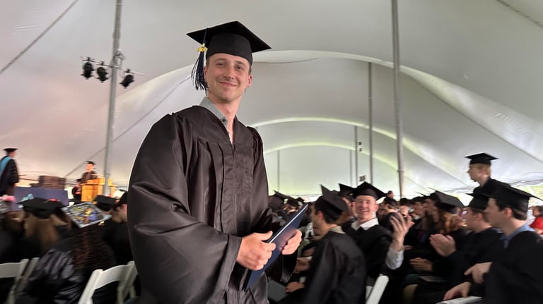 A student smiles and holds a diploma and wears a cap and gown at his graduation ceremony. 