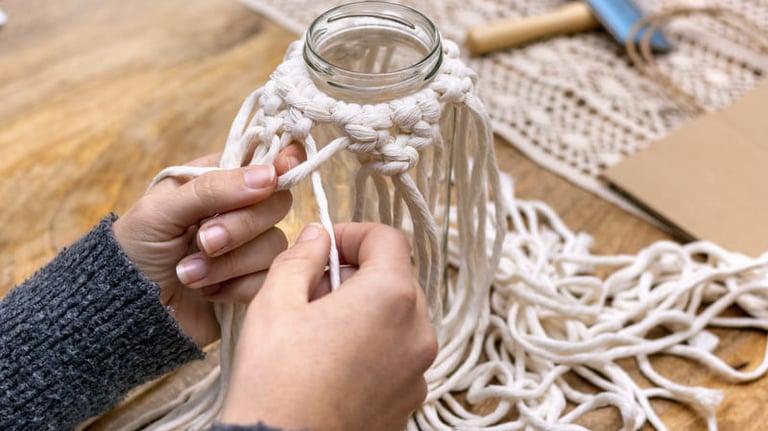 Manos haciendo nudos para un proyecto de macramé DIY alrededor de un frasco de vidrio sobre una mesa