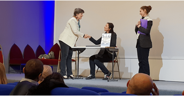 Sur scène une femme debout serre la main sous le regard d'une troisème forum accueil usager