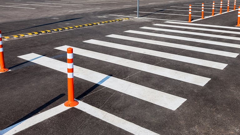 Painted white pedestrian crosswalk on asphalt with orange safety bollards in a parking lot.