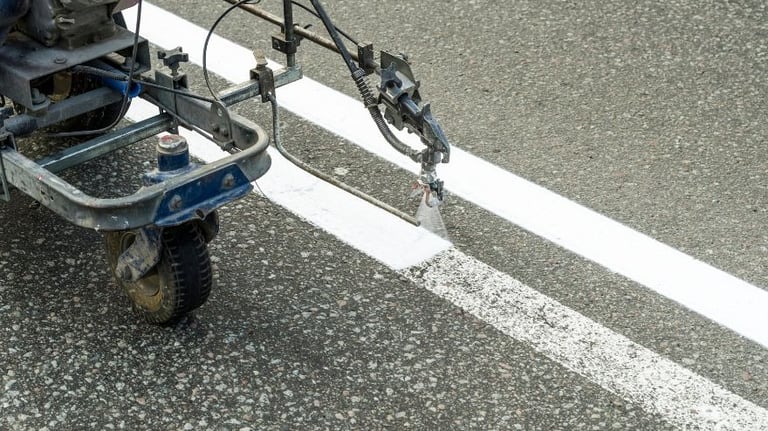 A road marking machine sprays white paint to create fresh stripes on an asphalt surface.
