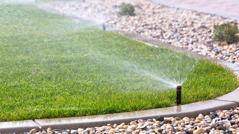 Automatic lawn sprinkler system watering a lush green backyard garden near a stone landscape border.