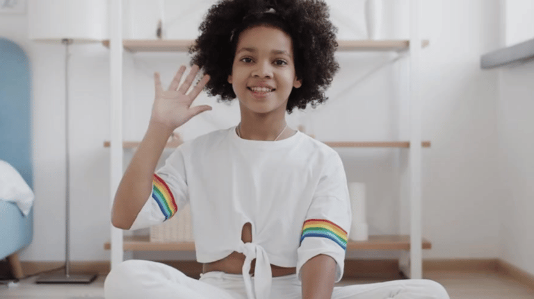 Little girl in a white pant set, sitting facing the camera waving hello
