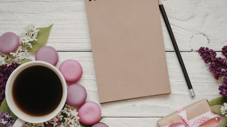 Flat lay of a blank kraft notebook with coffee, purple macarons, and lilac flowers on a white wood desk.