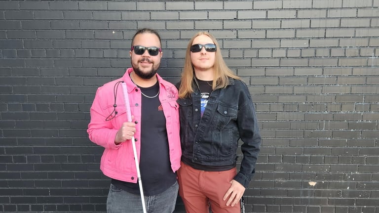 Two men in sunglasses posing against a black brick wall, one holding a white cane for the blind.