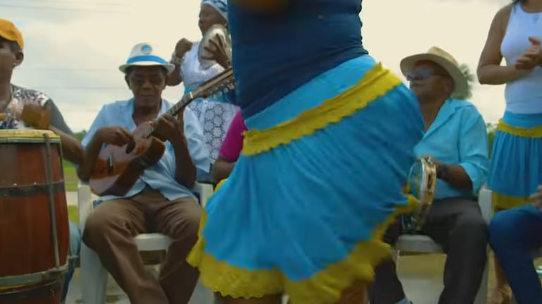 Grupo de samba de roda ao ar livre, com uma sambadeira vestida de azul ao centro e músicos ao redor.