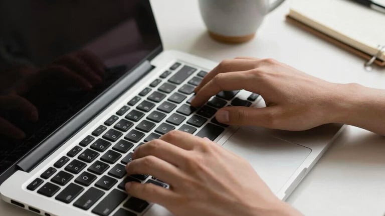 Detail of hands typing on a sleek, silver laptop keyboard. Beside the laptop is a small ceramic cup of coffee and a notebook. Minimalist desk setup with soft natural light in a professional study.