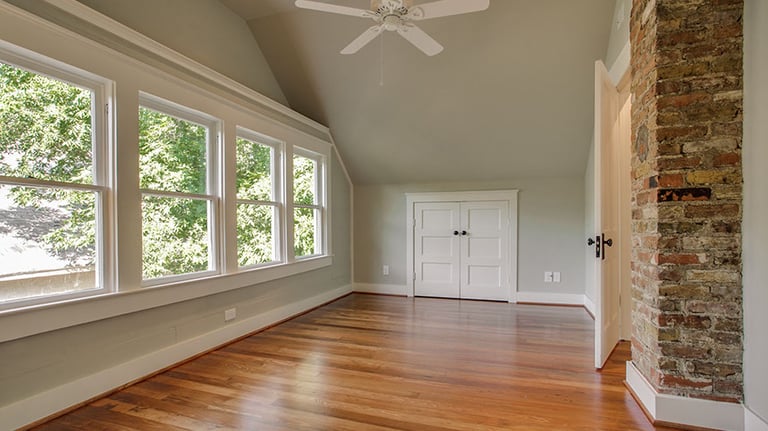 Empty modern attic bedroom with hardwood floors, exposed brick chimney, and large windows.
