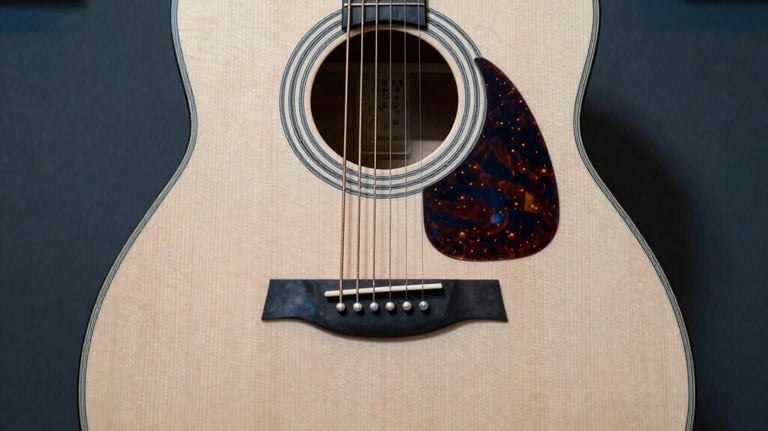 A top-down view of a modern acoustic guitar in a professional music studio, with soft light hitting the off-white wood grain and blue shadows.