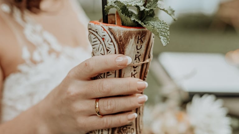 A bride in a white wedding dress holds a tropical tiki cocktail mug garnished with fresh mint.