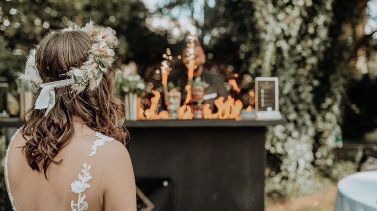 A bride with a floral crown watches a flair bartender perform a fire show at an outdoor wedding reception.