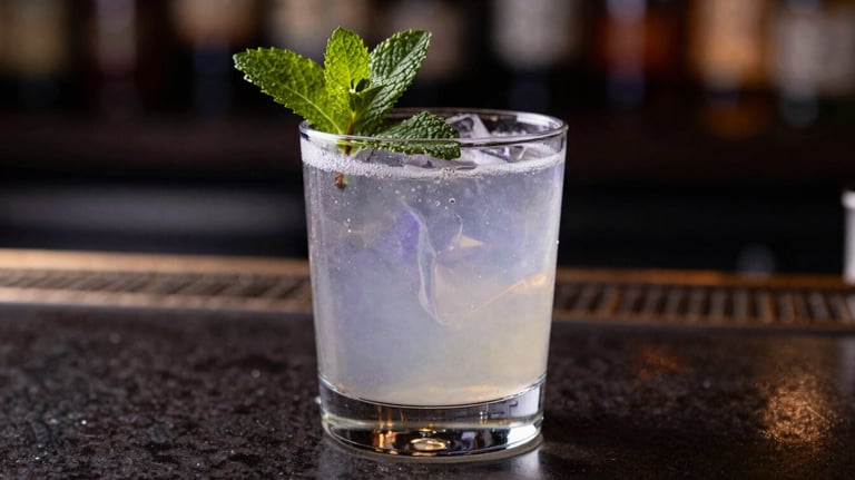 A close-up of a sophisticated cocktail garnished with fresh mint, sitting on a dark polished bar. The liquid reflects soft lavender gray and deep midnight indigo lights.
