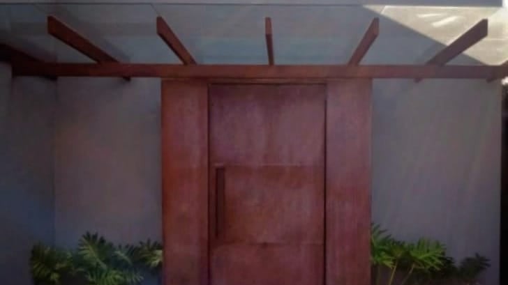 Porta de entrada pivotante de grandes dimensões, com revestimento que imita a aparência enferrujada do aço corten, sob uma pé