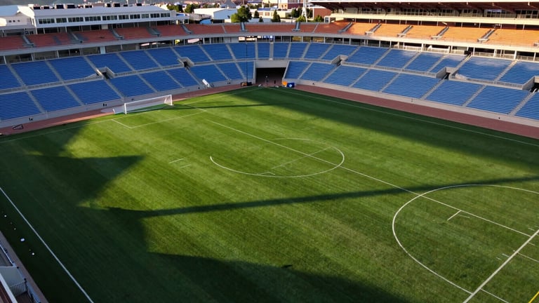 High-contrast aerial drone photography of a modern sports park in Eagle Mountain Utah, sharp white field markings on vibrant green turf, dramatic late afternoon sun casting long shadows, 35mm