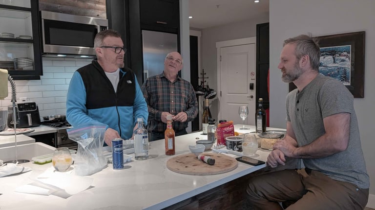 Three men talking around a kitchen island with drinks and food during a casual evening at home.