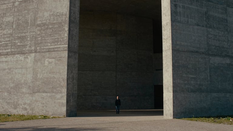 Cinematic film photography, moody natural light, high contrast, desaturated earth tones, wide anamorphic frame, 35mm grain, extreme wide shot of a colossal brutalist concrete structure with a tiny human figure standing in the shadows