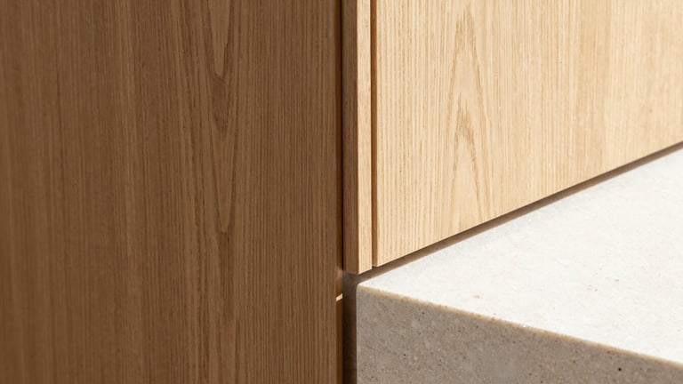 Extreme close-up of a seamless junction between warm rift-sawn oak wood paneling and a cool, smooth Belgian bluestone slab. Bright natural light highlights the textures.