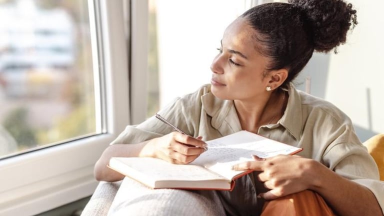 Thoughtful young woman journaling in a notebook by a window with natural light.
