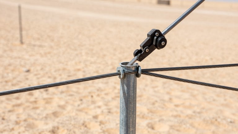 Close-up of a heavy-duty steel tension wire being tightened on a galvanized fence post by industrial tools, desert dust, bright sunlight