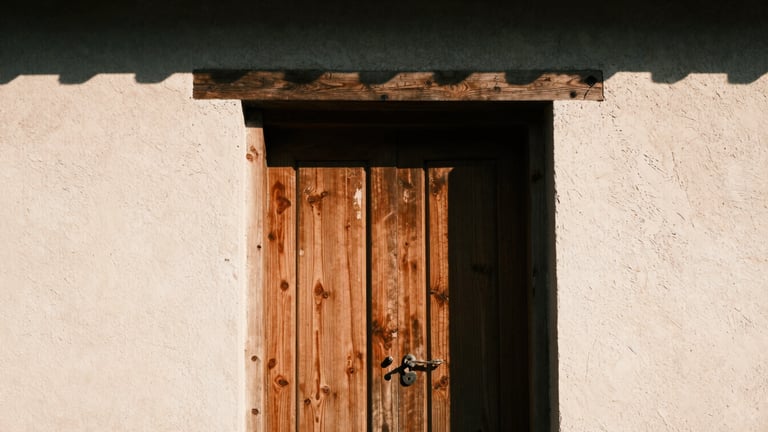 Tight abstract crop of a weathered teak door frame set against raw textured plaster, harsh high-contrast sunlight casting deep shadows, 35mm film.