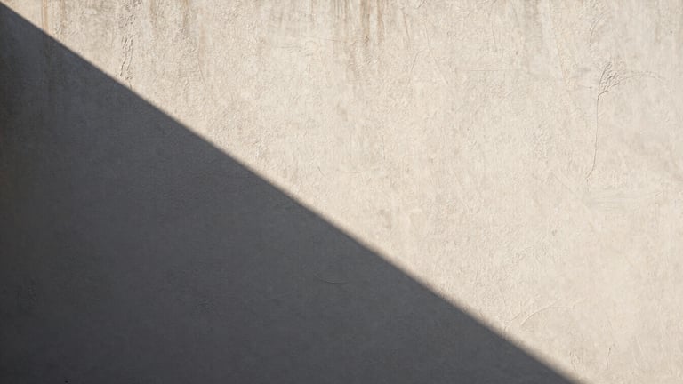 Wide bleed image of a textured plaster wall under harsh direct sunlight, creating a deep, sharp geometric shadow.